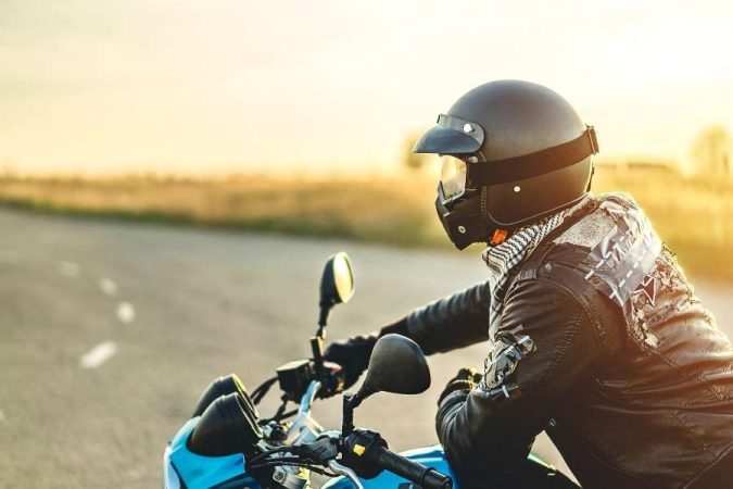 Un motard en veste de cuir et casque noir sur une moto bleue, roulant au coucher du soleil pour réduire la consommation de carburant.