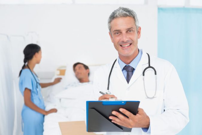 Un médecin de garde souriant aux cheveux gris, en blouse blanche avec un stéthoscope, écrivant sur un dossier dans une chambre d'hôpital, avec une infirmière et un patient flous en arrière-plan