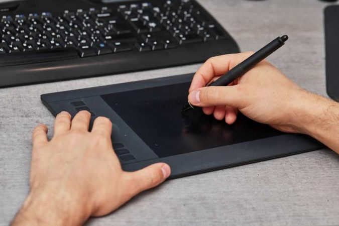 Gros plan sur les mains d'une personne utilisant un stylet sur une tablette graphique noire, posée sur un bureau gris près d'un clavier.