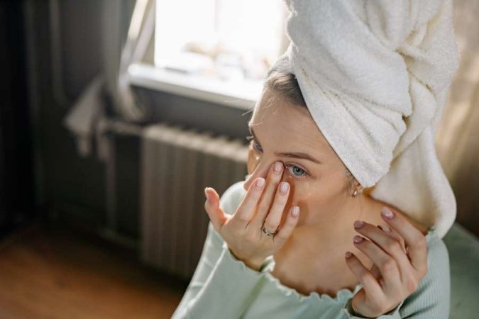 Une jeune femme avec une serviette sur la tête applique délicatement un soin sous ses yeux pour atténuer les cernes.