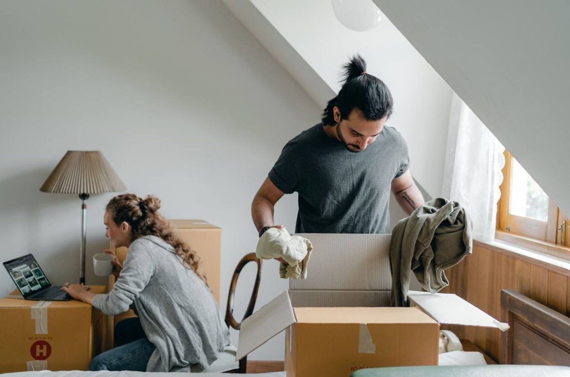 Un couple prépare ses cartons dans un appartement mansardé, l'homme pliant des vêtements tandis que la femme utilise un ordinateur pour tester un simulateur de devis de déménagement en ligne gratuit et rapide.
