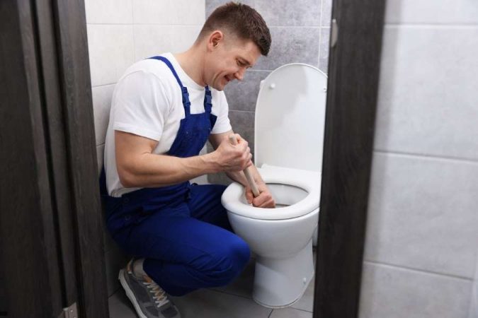 Un plombier en bleu de travail utilise une ventouse pour déboucher un WC dans une salle de bain carrelée.